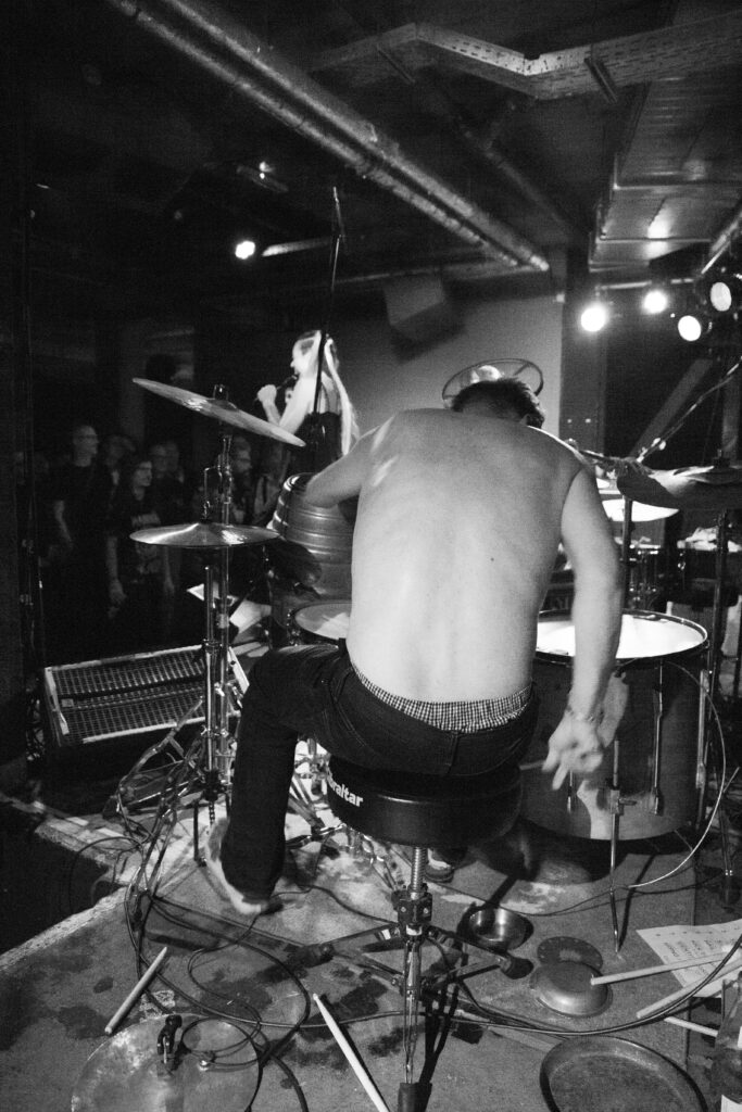 The Drummer of Maschin Kaput playing a gig at Hafenklang, Hamburg. A gritty black and white picture with a bare back and a hint of the audience